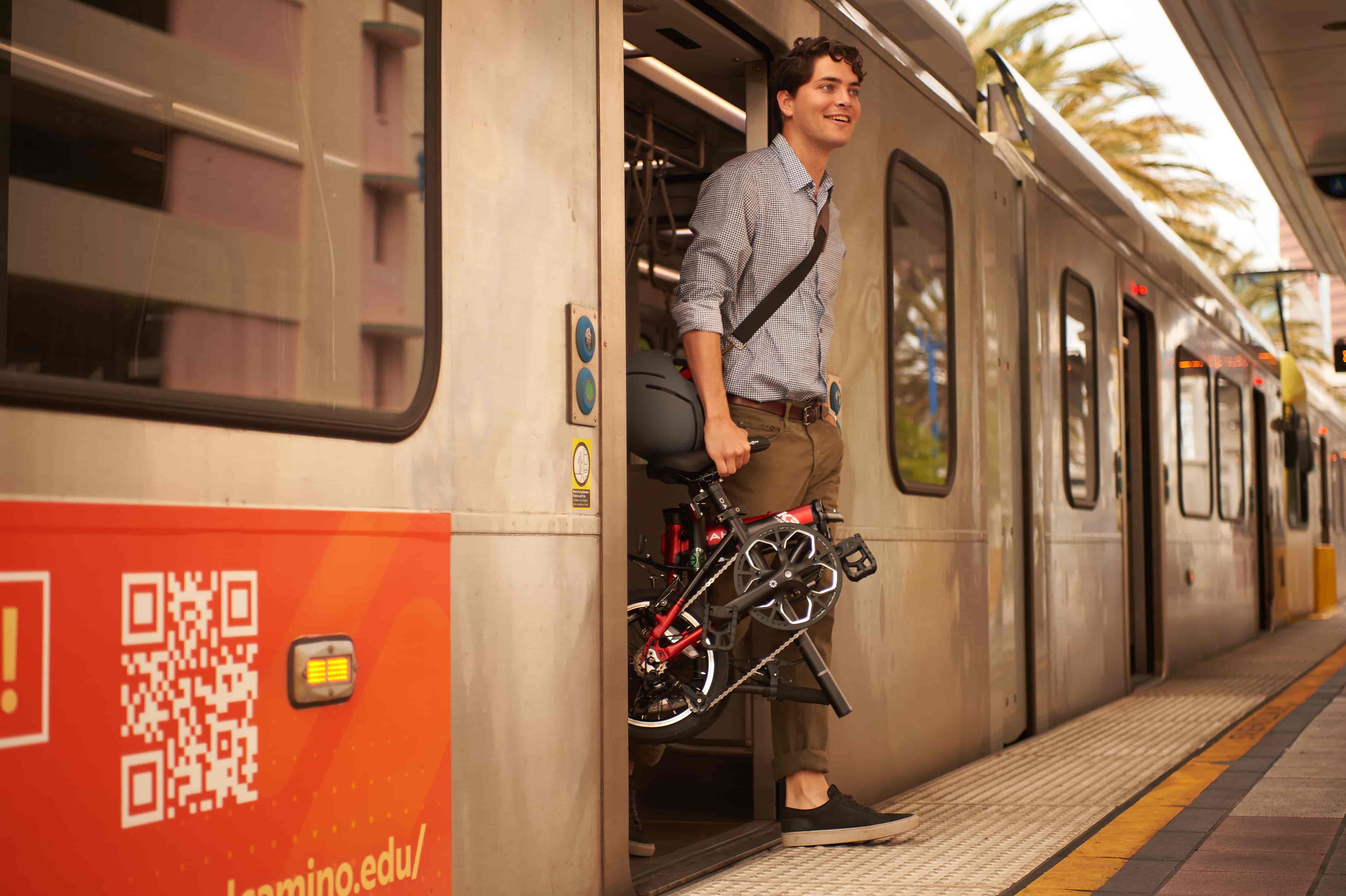 A man is stepping off a train while carrying a folded bicycle. He is dressed in casual attire and appears to be at a subway or train station platform.