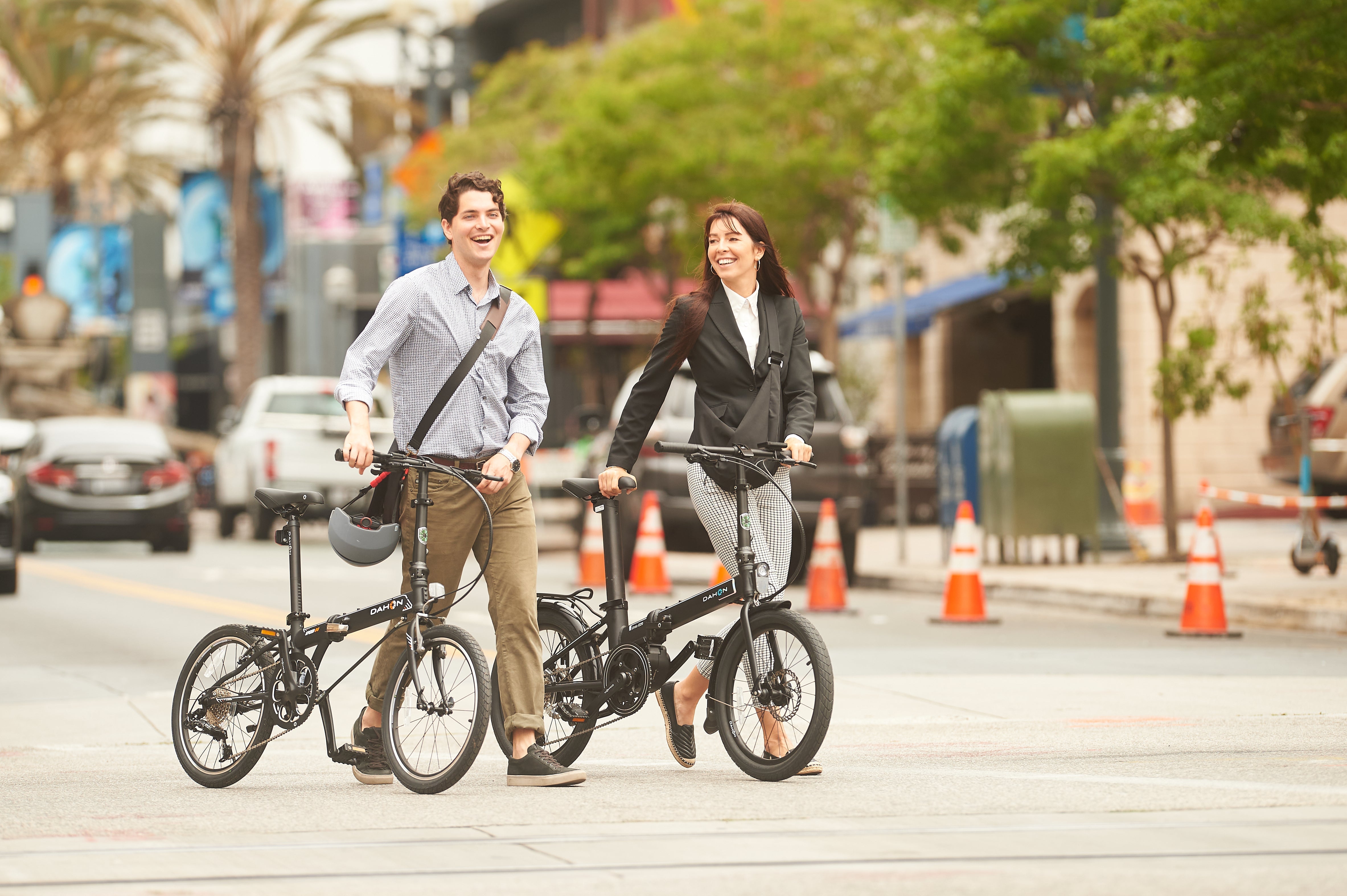 A man and a woman are walking on a city street, each carrying a folded bicycle. They appear to be in conversation, with cars and traffic cones visible in the background.